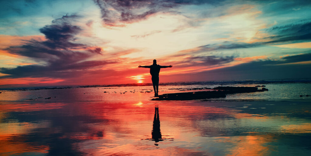 Person står med utsträckta armar i solnedgång vid havet